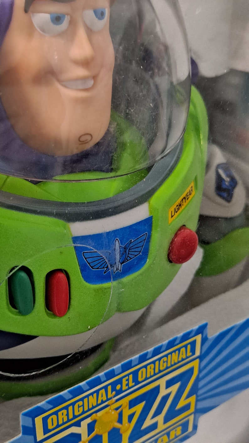 May include: A green and white Buzz Lightyear toy with a blue space ranger emblem on the chest. The toy is in a clear plastic package with the words "ORIGINAL - EL ORIGINAL" and "Buzz" printed on the bottom. The toy has a red button on the side and a yellow label that says "LIGHTYEAR".