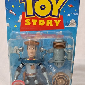May include: A plastic action figure of Woody from Toy Story, dressed in a space suit with a silver helmet and a brown cowboy hat. He is holding a silver and gold space blaster. The packaging is blue and red with stars and the words "Disney's Toy Story" and "Space Sheriff Woody Firing Turbo Blaster!"