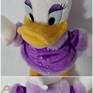 May include: A plush toy of Daisy Duck wearing a purple dress with a white fur trim and a purple bow in her hair.