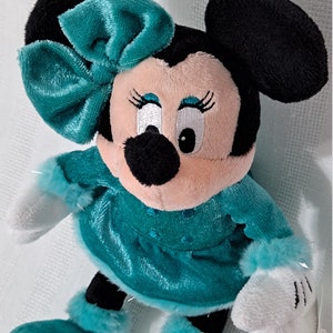 May include: A plush toy of Minnie Mouse wearing a teal blue dress with white fur trim and a matching teal blue bow. She has black ears and a black nose.