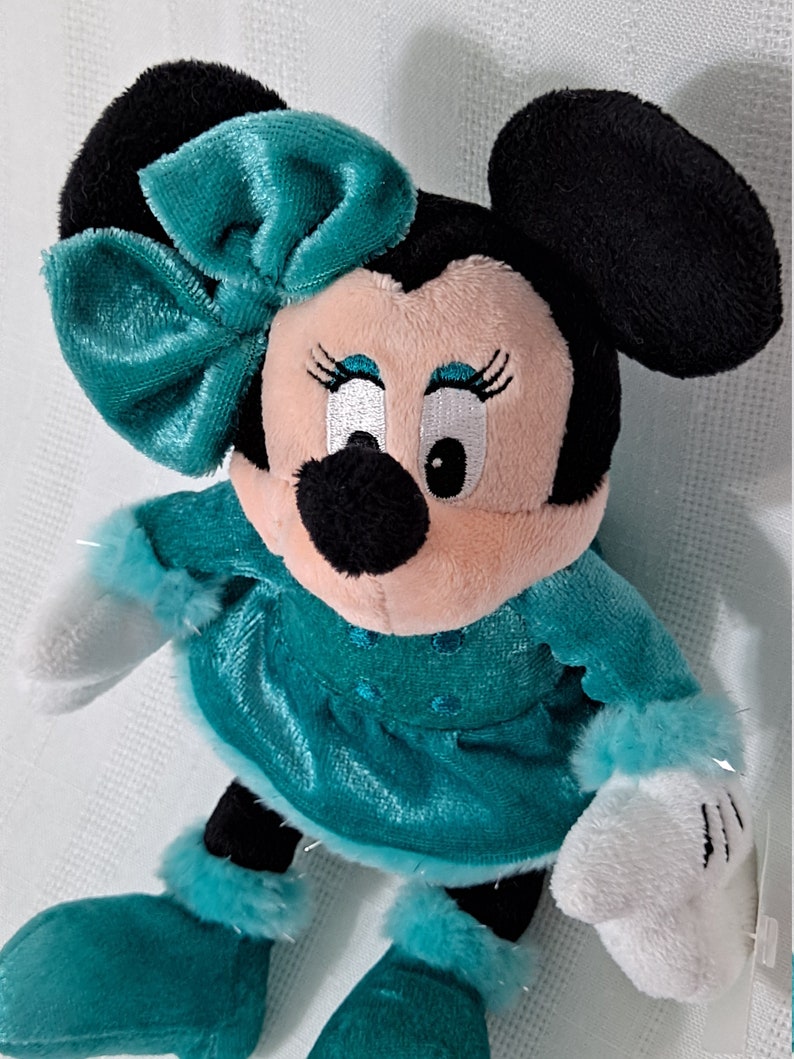 May include: A plush toy of Minnie Mouse wearing a teal blue dress with white fur trim and a matching teal blue bow. She has black ears and a black nose.