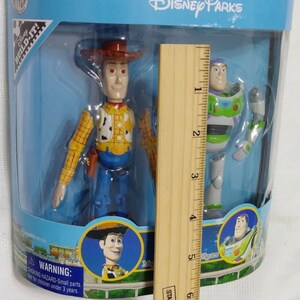 May include: A plastic box with a blue background and the text "Disney Parks" on the top. Inside the box are two action figures: Woody from Toy Story, wearing his signature cowboy hat and vest, and Buzz Lightyear, wearing his space ranger suit. The figures are approximately 6 inches tall. The box also includes a warning label about choking hazards for children under 3 years old.