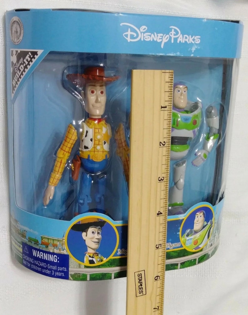 May include: A plastic box with a blue background and the text "Disney Parks" on the top. Inside the box are two action figures: Woody from Toy Story, wearing his signature cowboy hat and vest, and Buzz Lightyear, wearing his space ranger suit. The figures are approximately 6 inches tall. The box also includes a warning label about choking hazards for children under 3 years old.