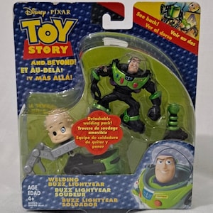 May include: Toy Story Buzz Lightyear action figure with a welding pack. The figure is green and black with a clear plastic helmet. The welding pack is green and silver. The packaging is red, blue, and yellow with the Toy Story logo and the words "And Beyond!" in English, French, and Spanish.