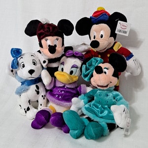 May include: Five plush Disney characters dressed in winter clothing. Mickey Mouse is wearing a red and gold uniform, Minnie Mouse is wearing a black and white striped dress, Daisy Duck is wearing a purple dress, Pluto is wearing a blue scarf, and Goofy is wearing a blue dress.