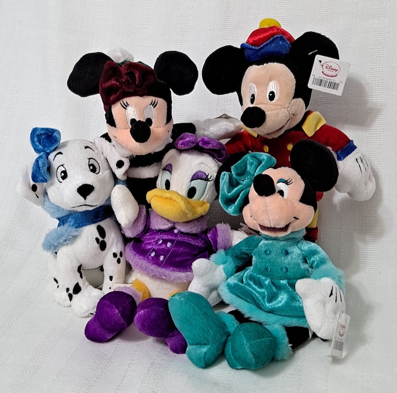 May include: Five plush Disney characters dressed in winter clothing. Mickey Mouse is wearing a red and gold uniform, Minnie Mouse is wearing a black and white striped dress, Daisy Duck is wearing a purple dress, Pluto is wearing a blue scarf, and Goofy is wearing a blue dress.