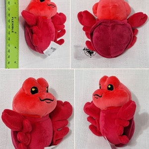May include: A red plush crab toy with a Disney logo tag. The crab has large, black eyes and a smiling mouth.