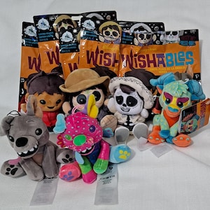 Puede incluir: Una colección de peluches Disney Wishables, con personajes de la serie Coco Plaza de la Familia. Los peluches incluyen un perro gris, una criatura rosa y verde y personajes con sombreros. El embalaje es naranja y negro.
