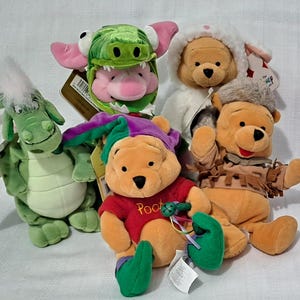 May include: A collection of plush Winnie the Pooh characters in various costumes. The plush toys include a green dragon, a pig in a crocodile hat, Pooh in a bunny outfit, and Pooh in a Native American-inspired outfit. The Pooh plush toys are wearing a red shirt with the word "Pooh".