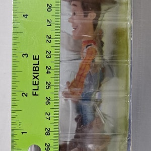May include: A plastic-wrapped toy figure of Woody from the Toy Story franchise. Woody is wearing his signature cowboy hat, vest, and jeans. The figure is approximately 15 cm tall.