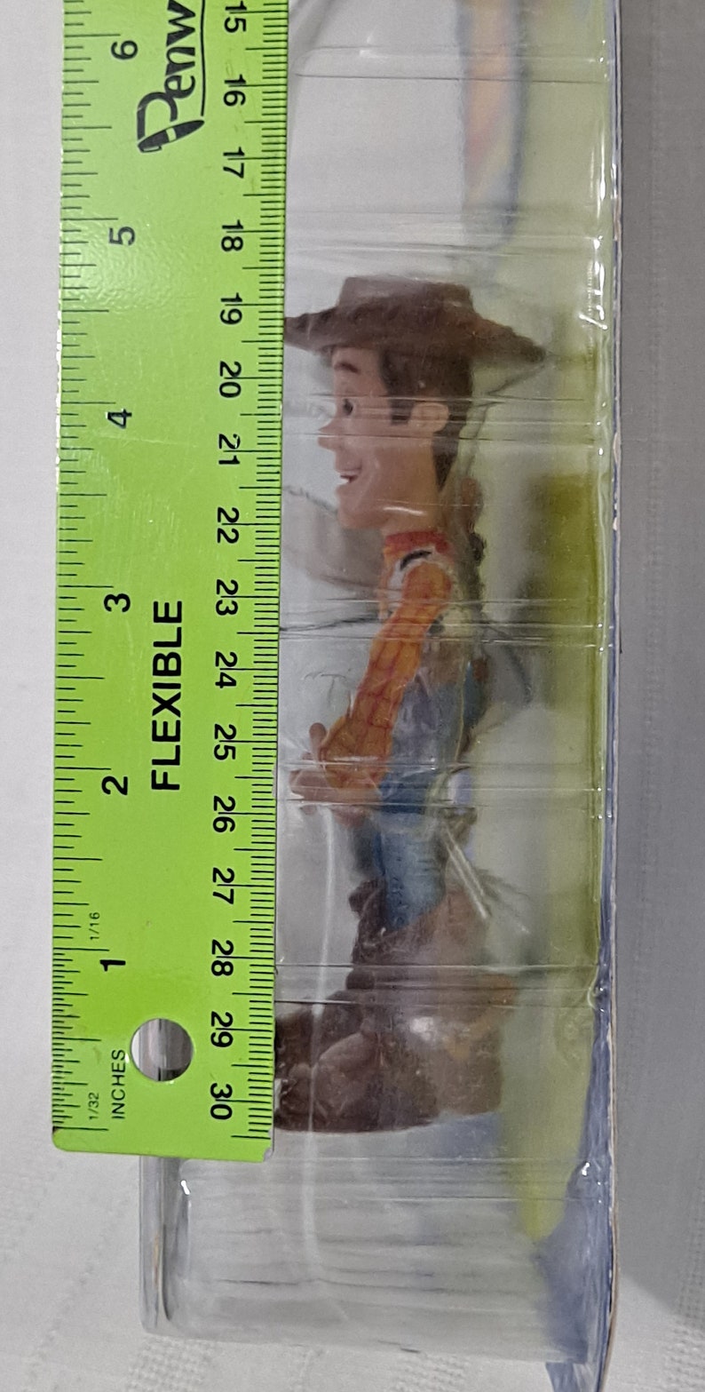 May include: A plastic-wrapped toy figure of Woody from the Toy Story franchise. Woody is wearing his signature cowboy hat, vest, and jeans. The figure is approximately 15 cm tall.