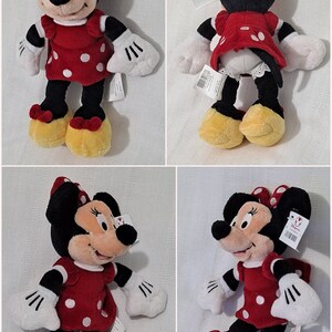 May include: A red and white polka dot dress with a red bow on top of the head. The plush toy is a black and white mouse with yellow shoes. The toy is wearing a white shirt with black cuffs. The toy is holding its hands up in the air. The toy is sitting on a white background.