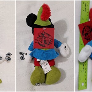 Op de afbeelding: Een pluche Mickey Mouse speelgoed dat een groen en rood Robin Hood kostuum draagt. Het speelgoed is ongeveer 30 cm hoog en heeft een label met de tekst "Mickey's Magical Adventure".