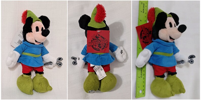 Op de afbeelding: Een pluche Mickey Mouse speelgoed dat een groen en rood Robin Hood kostuum draagt. Het speelgoed is ongeveer 30 cm hoog en heeft een label met de tekst "Mickey's Magical Adventure".