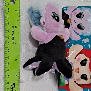 May include: A purple and blue plush toy of Ursula from The Little Mermaid. The toy is about 5 inches tall and has a black octopus tail with white polka dots. The toy is packaged in a plastic bag with a picture of Ursula and Ariel on it.