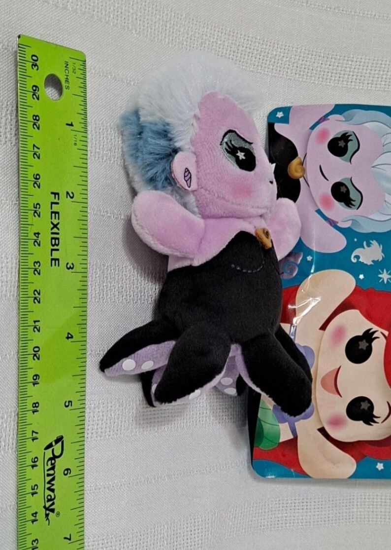 May include: A purple and blue plush toy of Ursula from The Little Mermaid. The toy is about 5 inches tall and has a black octopus tail with white polka dots. The toy is packaged in a plastic bag with a picture of Ursula and Ariel on it.