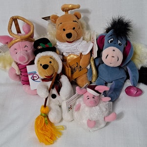 May include: Five plush Disney characters dressed as angels.  The characters are Winnie the Pooh, Piglet, Eeyore, and two Piglets.  The characters are all wearing angel wings and halos.  The Winnie the Pooh character is holding a music book and a golden trumpet.  The Piglet character is wearing a pink and white striped dress.  The Eeyore character is wearing a blue and purple dress.  The two Piglet characters are wearing white dresses with pink accents.