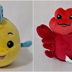 May include: Two plush toys, a yellow fish with blue fins and a red lobster. Both toys have big eyes with glitter accents and smiling mouths.