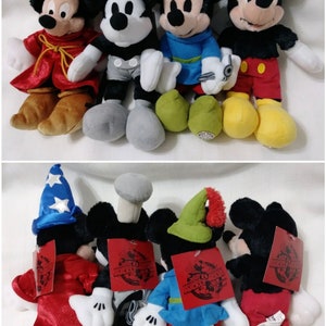 Op de afbeelding: Vier pluche Mickey Mouse-speelgoedjes gekleed in verschillende kostuums. De eerste is een tovenaar met een blauwe hoed en een rode mantel. De tweede is een Steamboat Willie met een zwart-witte outfit. De derde is een Robin Hood met een groene hoed en een blauw shirt. De vierde is een piraat met een zwart ooglapje en een rode broek. Alle vier speelgoedjes hebben een Mickey Mouse-label op hun rug met de tekst "Celebrating 90 Years of Mickey Mouse".