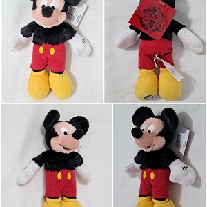 Op de afbeelding: Een zwart en rood pluche Mickey Mouse speelgoed met gele schoenen. Het speelgoed draagt een rood shirt met twee gele knopen en een zwarte broek. Het speelgoed heeft een label met de tekst "Celebrating 90 Years with Mickey Mouse".
