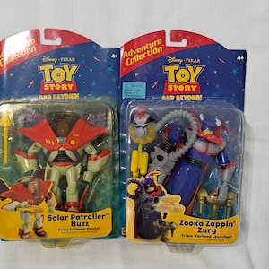 May include: Two action figures from the Disney Pixar Toy Story franchise. The figures are packaged in plastic blister packs with cardboard backing. The figure on the left is Solar Patroller Buzz, a white and red robot with a yellow missile. The figure on the right is Zooka Zappin' Zurg, a blue and red robot with a yellow triple warhead launcher. Both figures are part of the Adventure Collection.