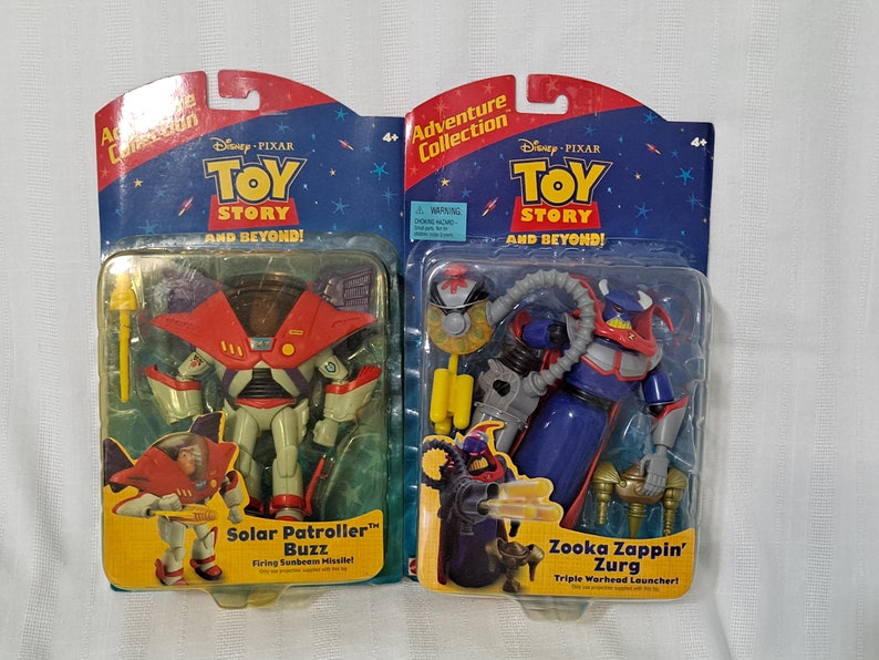 May include: Two action figures from the Disney Pixar Toy Story franchise. The figures are packaged in plastic blister packs with cardboard backing. The figure on the left is Solar Patroller Buzz, a white and red robot with a yellow missile. The figure on the right is Zooka Zappin' Zurg, a blue and red robot with a yellow triple warhead launcher. Both figures are part of the Adventure Collection.