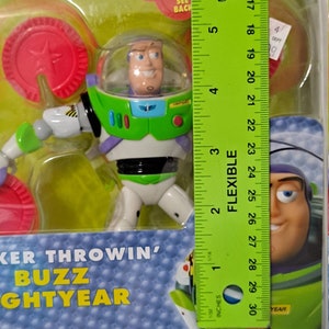 May include: A plastic action figure of Buzz Lightyear from the Toy Story franchise. The figure is wearing his signature green and white space ranger suit. The figure is holding a red disc in his right hand. The packaging includes the text "Checker Throwin' Buzz Lightyear" and "Ages 4+".