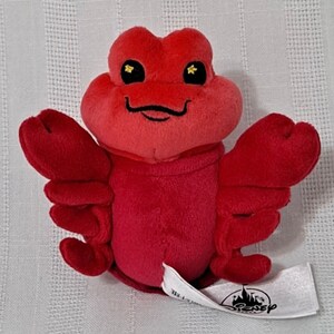May include: Red plush toy of Sebastian the crab from Disney's The Little Mermaid. He has a big smile and gold eyes. The toy has a Disney tag attached to one of his claws.