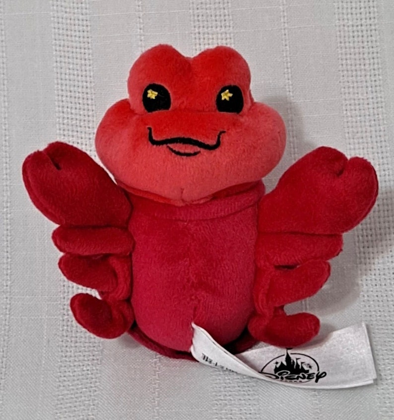 May include: Red plush toy of Sebastian the crab from Disney's The Little Mermaid. He has a big smile and gold eyes. The toy has a Disney tag attached to one of his claws.