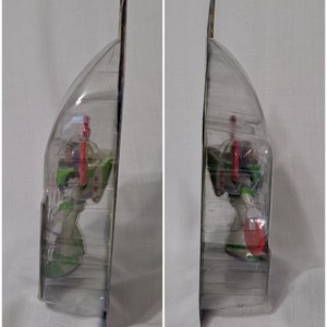 May include: A plastic-wrapped action figure of Buzz Lightyear from the Toy Story franchise. The figure is in a standing pose with his arms outstretched and his head tilted slightly to the side. The figure is wearing his signature space ranger suit, which is green, white, and purple. The figure is holding a red laser gun in his right hand. The figure is packaged in a clear plastic blister pack with a blue and yellow cardboard backing.