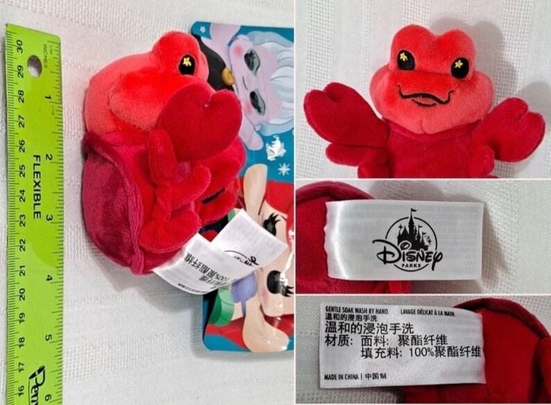 May include: Red plush crab toy with a smiling face and large, black eyes. The toy is approximately 4 inches tall and is labeled "Disney Parks".