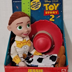 K&ouml;nnte beinhalten: Ein Pl&uuml;schtier von Jessie aus Toy Story 2. Jessie tr&auml;gt ihr charakteristisches Outfit, ein wei&szlig;es Shirt mit gelbem Besatz, eine braune Weste, eine blaue Jeans mit einem Kuhflecken-Patch und braune Stiefel. Sie sitzt mit ihrem roten Cowboyhut neben sich. Die Verpackung zeigt das Toy Story 2 Logo und den Text "Die perfekte Gr&ouml;&szlig;e zum Kuscheln!"