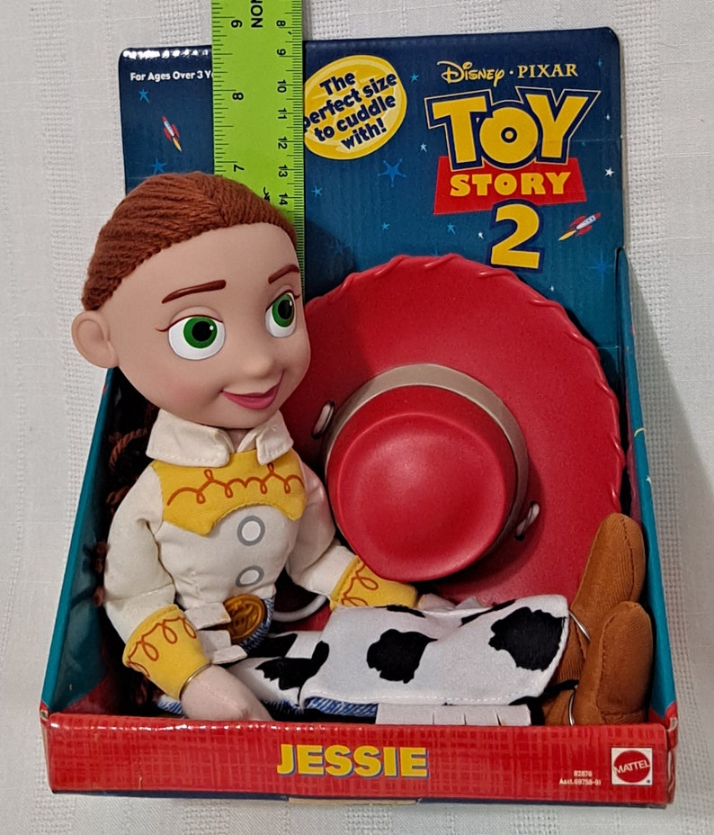 K&ouml;nnte beinhalten: Ein Pl&uuml;schtier von Jessie aus Toy Story 2. Jessie tr&auml;gt ihr charakteristisches Outfit, ein wei&szlig;es Shirt mit gelbem Besatz, eine braune Weste, eine blaue Jeans mit einem Kuhflecken-Patch und braune Stiefel. Sie sitzt mit ihrem roten Cowboyhut neben sich. Die Verpackung zeigt das Toy Story 2 Logo und den Text "Die perfekte Gr&ouml;&szlig;e zum Kuscheln!"
