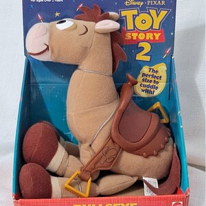 K&ouml;nnte beinhalten: Ein braunes Pl&uuml;schpferd mit Sattel und gelbem Gurt. Das Pferd sitzt auf einem roten und blauen Karton mit den Worten "Disney Pixar Toy Story 2" und "Bullseye" darauf gedruckt. Der Karton sagt auch "Die perfekte Gr&ouml;&szlig;e zum Kuscheln!" und "F&uuml;r Kinder ab 3 Jahren".