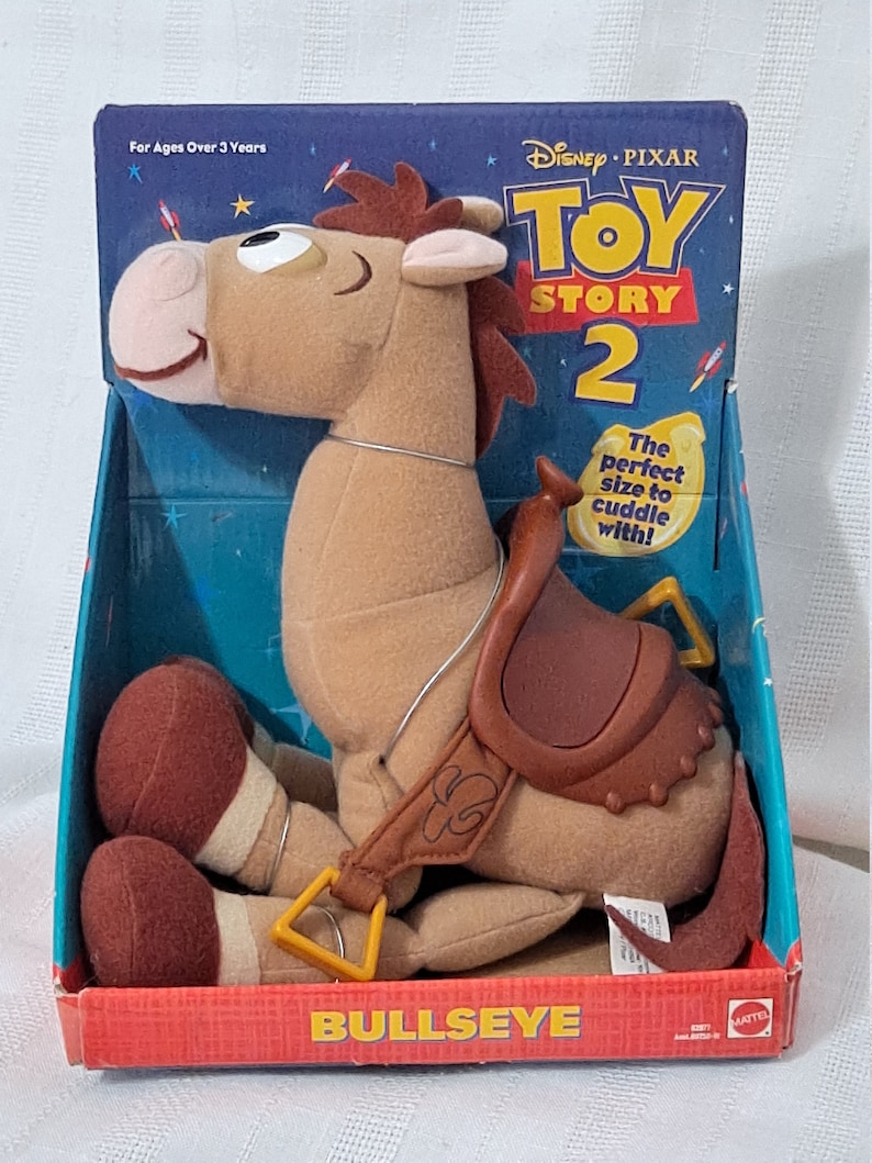 K&ouml;nnte beinhalten: Ein braunes Pl&uuml;schpferd mit Sattel und gelbem Gurt. Das Pferd sitzt auf einem roten und blauen Karton mit den Worten "Disney Pixar Toy Story 2" und "Bullseye" darauf gedruckt. Der Karton sagt auch "Die perfekte Gr&ouml;&szlig;e zum Kuscheln!" und "F&uuml;r Kinder ab 3 Jahren".