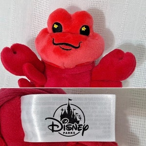 May include: Red plush lobster toy with black eyes and a black smile. The toy has a white tag attached with the Disney Parks logo.
