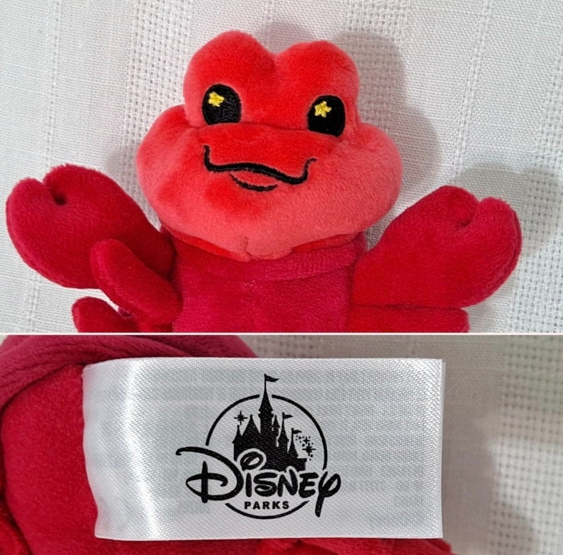 May include: Red plush lobster toy with black eyes and a black smile. The toy has a white tag attached with the Disney Parks logo.