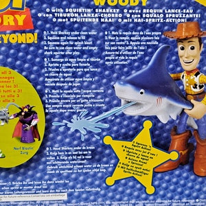May include: Woody from Toy Story action figure riding a blue shark water squirter toy. The shark has a white mouth and teeth. The toy is labelled with "Squirtin' Sharky" and has instructions for use in multiple languages.