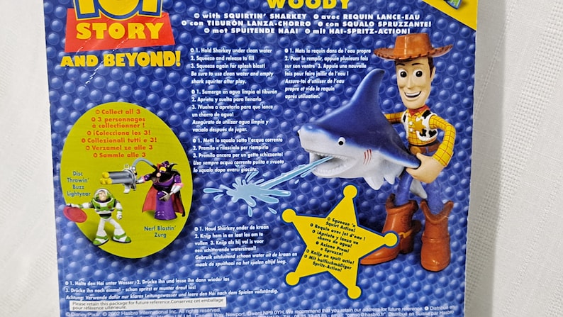 May include: Woody from Toy Story action figure riding a blue shark water squirter toy. The shark has a white mouth and teeth. The toy is labelled with "Squirtin' Sharky" and has instructions for use in multiple languages.