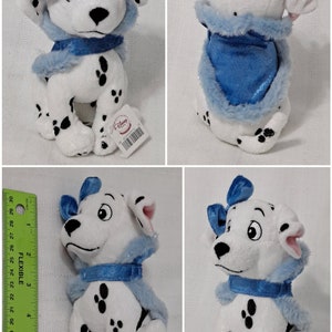 May include: A white and black Dalmatian plush toy wearing a blue and white fur-trimmed scarf and a blue bow. The plush toy is sitting with its legs tucked underneath it. The toy has a Disney Store tag attached to it.