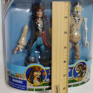 May include: A plastic toy set featuring Captain Jack Sparrow and the skeleton from Pirates of the Caribbean. The set is packaged in a blue and white box with the Disney Parks logo. The box includes a ruler for size reference.