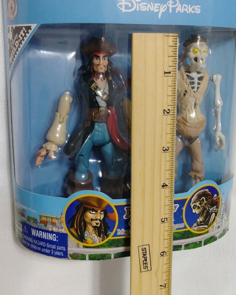May include: A plastic toy set featuring Captain Jack Sparrow and the skeleton from Pirates of the Caribbean. The set is packaged in a blue and white box with the Disney Parks logo. The box includes a ruler for size reference.