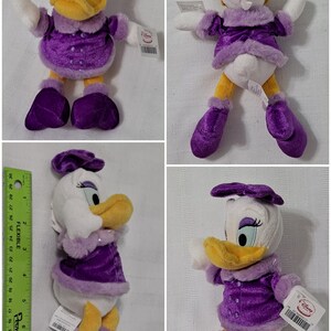 May include: A plush toy of Daisy Duck wearing a purple dress with a white fur trim, a purple bow, and purple boots. The toy is sitting on a white background.