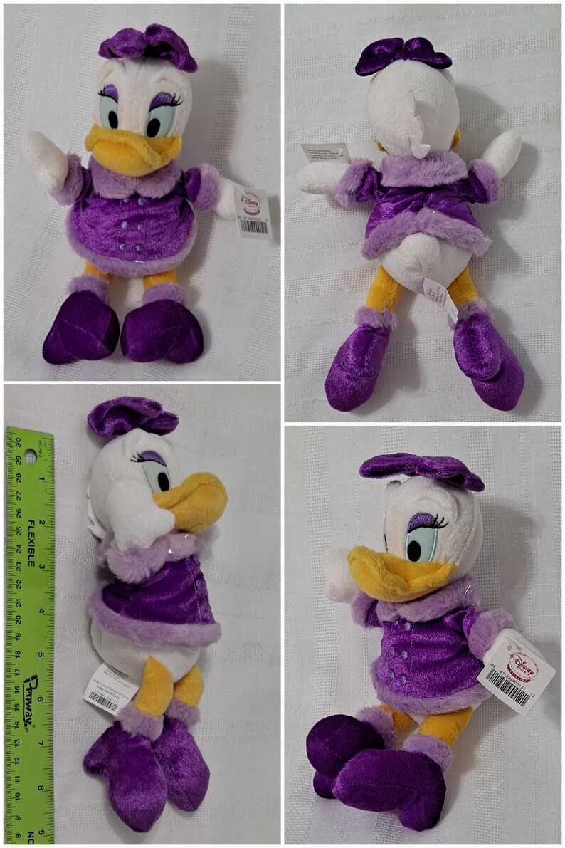 May include: A plush toy of Daisy Duck wearing a purple dress with a white fur trim, a purple bow, and purple boots. The toy is sitting on a white background.