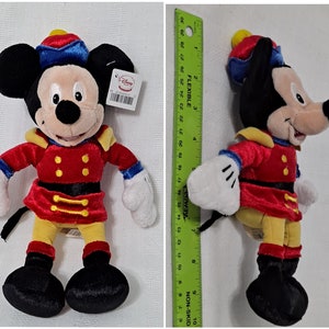 May include: A plush Mickey Mouse toy dressed as a nutcracker. He is wearing a red and gold uniform with a blue hat and a yellow sash. The toy is about 10 inches tall.
