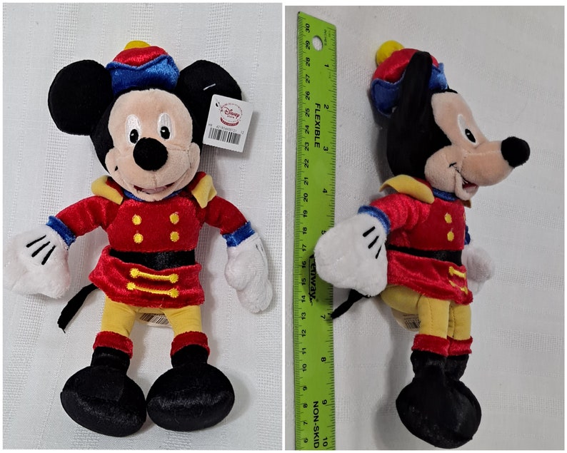 May include: A plush Mickey Mouse toy dressed as a nutcracker. He is wearing a red and gold uniform with a blue hat and a yellow sash. The toy is about 10 inches tall.