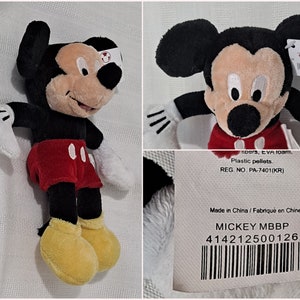 May include: A black and red Mickey Mouse plush toy with yellow shoes. The toy is approximately 10 inches tall and has a white tag attached to its right ear. The tag features the Disney logo and the text "MICKEY MBBP 414212500126".