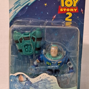 Peut inclure: Une figurine d'action en plastique bleue et verte de Buzz l'&Eacute;clair de Toy Story 2. La figurine tient un pistolet &agrave; eau et porte un sac &agrave; dos. L'emballage est bleu et vert avec les mots "Aqua Action" et "Super Squirtin' Buzz" dessus.