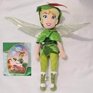 May include: A plush Tinker Bell doll with blonde hair, blue eyes, and a smiling expression. She wears a green leaf-shaped outfit, leggings, and shoes. The doll has translucent wings and a tag with the Disney Tinker Bell logo.