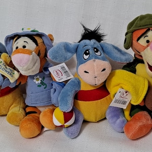 May include: Four plush Winnie the Pooh characters: Winnie the Pooh, Tigger, Eeyore, and Tigger. Winnie the Pooh is wearing a blue hat and holding a honey pot. Tigger is wearing a blue jacket and holding a ball. Eeyore is wearing a yellow shirt and red stripes. Tigger is wearing a green hat and holding a brown football.